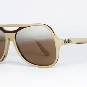 May include: Beige and brown aviator sunglasses with gradient lenses. The sunglasses have a thick frame with a bridge and arms. The Ray-Ban logo is visible on the right arm.