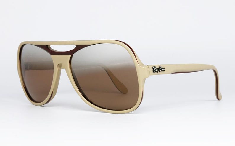 May include: Beige and brown aviator sunglasses with gradient lenses. The sunglasses have a thick frame with a bridge and arms. The Ray-Ban logo is visible on the right arm.