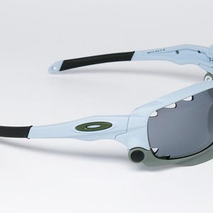 May include: Light blue and gray sports sunglasses with black accents. The sunglasses have a wraparound design with dark gray lenses and a green logo on the frame. The arms are black and light blue.