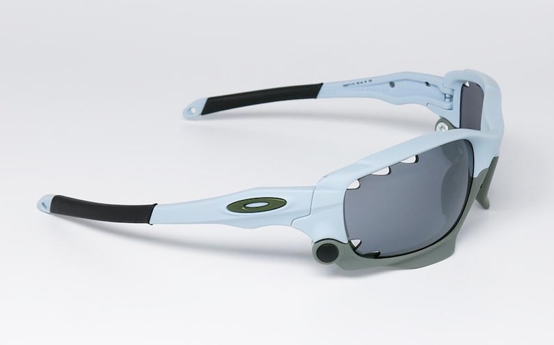 May include: Light blue and gray sports sunglasses with black accents. The sunglasses have a wraparound design with dark gray lenses and a green logo on the frame. The arms are black and light blue.