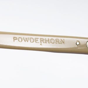 May include: Close-up of a tan-colored eyeglass arm with the word "POWDERHORN" embossed on the side. The arm is connected to the frame with a silver hinge.
