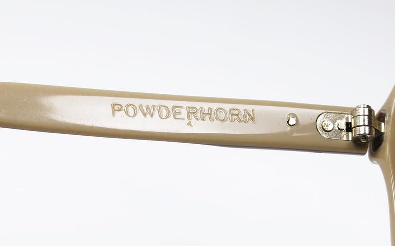 May include: Close-up of a tan-colored eyeglass arm with the word "POWDERHORN" embossed on the side. The arm is connected to the frame with a silver hinge.