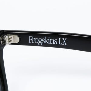 May include: Close-up of black sunglasses with the text "Frogskins LX" printed in white on the temple. The sunglasses have a silver hinge and dark lenses, set against a white background.