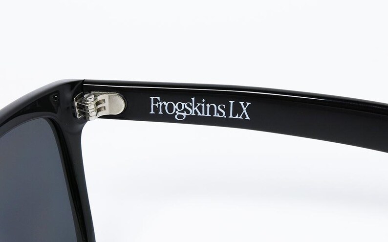 May include: Close-up of black sunglasses with the text "Frogskins LX" printed in white on the temple. The sunglasses have a silver hinge and dark lenses, set against a white background.