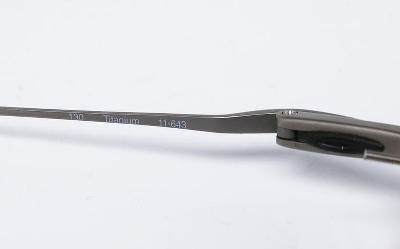 May include: Close-up of the arm of a pair of glasses. The arm is made of titanium and has the numbers 130, 11-643 printed on it.