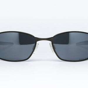 Może przedstawiać: Owalne okulary przeciwsłoneczne z czarną metalową oprawką i ciemnoszarymi soczewkami. Okulary mają przezroczyste noski. Design jest elegancki i nowoczesny, odpowiedni do r&oacute;żnych aktywności na świeżym powietrzu.