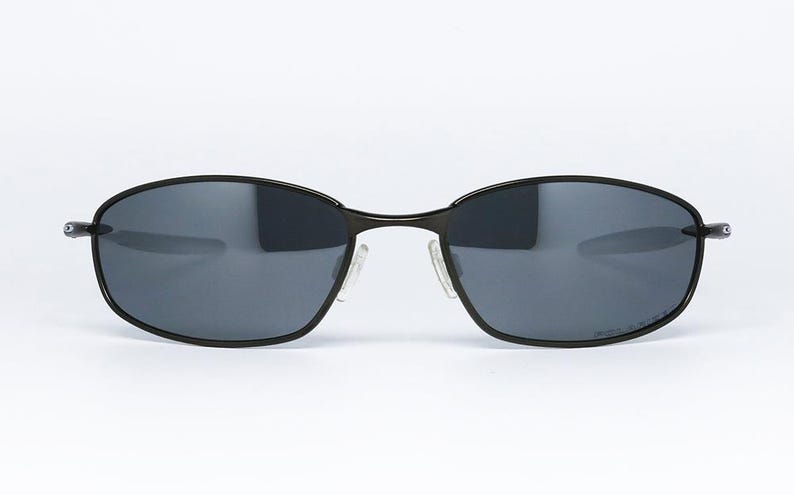 Może przedstawiać: Owalne okulary przeciwsłoneczne z czarną metalową oprawką i ciemnoszarymi soczewkami. Okulary mają przezroczyste noski. Design jest elegancki i nowoczesny, odpowiedni do r&oacute;żnych aktywności na świeżym powietrzu.