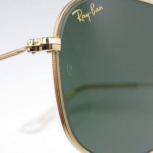 May include: Close-up of gold-tone Ray-Ban sunglasses with green lenses. The sunglasses have a subtle, textured frame and the Ray-Ban logo is visible on the right lens.