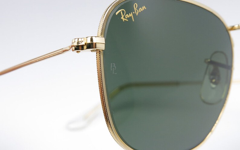 May include: Close-up of gold-tone Ray-Ban sunglasses with green lenses. The sunglasses have a subtle, textured frame and the Ray-Ban logo is visible on the right lens.