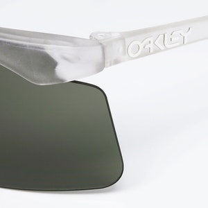 Puede incluir: Primer plano de una gafas de sol transparentes y semitransparentes con lentes verde oscuro. Las gafas de sol tienen un logotipo "Oakley" blanco en la patilla derecha.