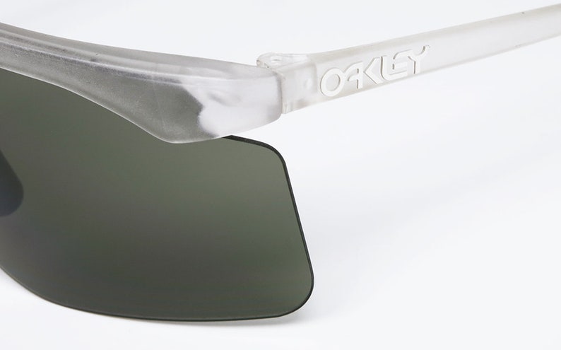 Puede incluir: Primer plano de una gafas de sol transparentes y semitransparentes con lentes verde oscuro. Las gafas de sol tienen un logotipo "Oakley" blanco en la patilla derecha.