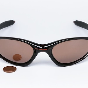Pode incluir: &Oacute;culos de sol pretos com lentes castanhas e o nome da marca "Oakley" em vermelho. Os &oacute;culos de sol t&ecirc;m um design curvo. Uma moeda &eacute; colocada na lente e outra moeda &eacute; colocada abaixo dos &oacute;culos.