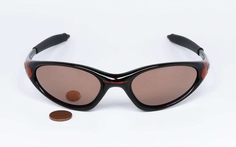 Pode incluir: &Oacute;culos de sol pretos com lentes castanhas e o nome da marca "Oakley" em vermelho. Os &oacute;culos de sol t&ecirc;m um design curvo. Uma moeda &eacute; colocada na lente e outra moeda &eacute; colocada abaixo dos &oacute;culos.