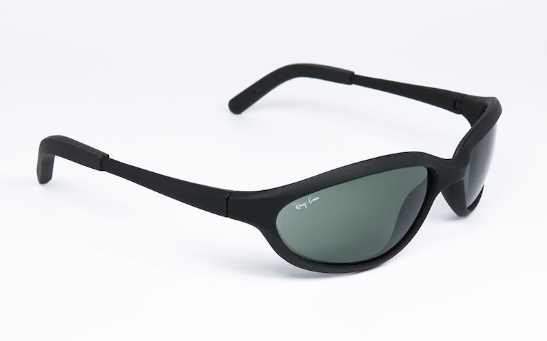 Pode incluir: &Oacute;culos de sol pretos com arma&ccedil;&atilde;o curva e lentes cinzentas. O log&oacute;tipo Ray-Ban &eacute; vis&iacute;vel na lente. Os &oacute;culos de sol t&ecirc;m um design desportivo e s&atilde;o adequados para atividades ao ar livre.