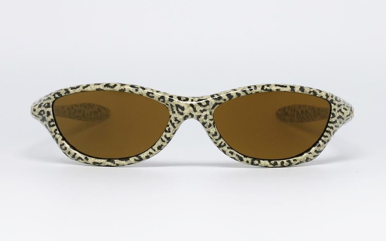 May include: Sunglasses with a leopard print pattern on the frames. The lenses are a warm brown color. The sunglasses are set against a white background.