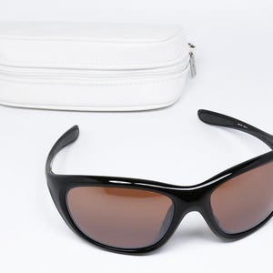 K&ouml;nnte beinhalten: Schwarze Sonnenbrille mit braunen Gl&auml;sern und einem wei&szlig;en Etui mit Rei&szlig;verschluss. Die Sonnenbrille hat ein schlankes, geschwungenes Design. Das Etui ist rechteckig und aus glattem, wei&szlig;em Material.