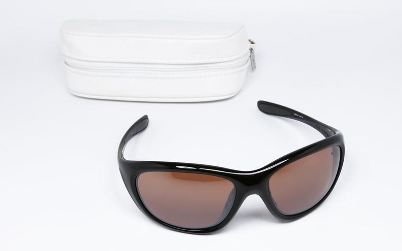 K&ouml;nnte beinhalten: Schwarze Sonnenbrille mit braunen Gl&auml;sern und einem wei&szlig;en Etui mit Rei&szlig;verschluss. Die Sonnenbrille hat ein schlankes, geschwungenes Design. Das Etui ist rechteckig und aus glattem, wei&szlig;em Material.
