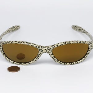 May include: Sunglasses with a leopard print pattern on the frames and arms. The lenses are a warm brown color. A penny is placed near the sunglasses for scale.