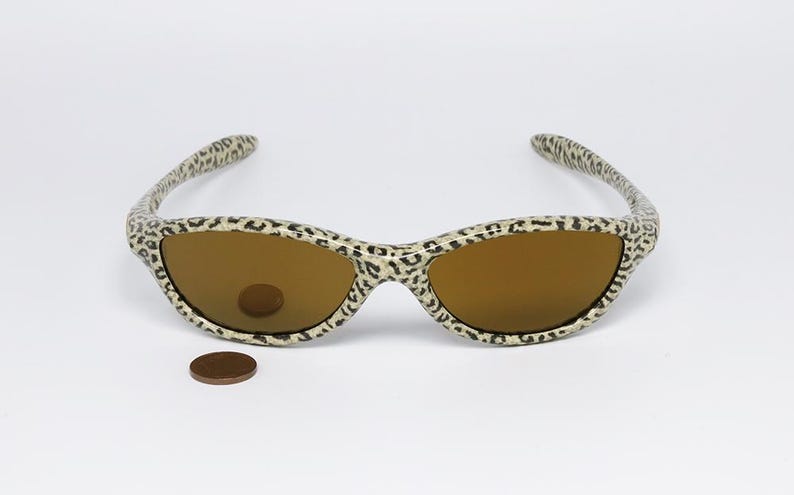 May include: Sunglasses with a leopard print pattern on the frames and arms. The lenses are a warm brown color. A penny is placed near the sunglasses for scale.