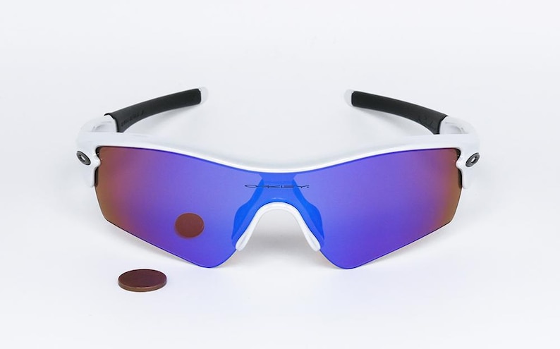 Pu&ograve; includere: Occhiali da sole con montatura bianca e lenti a specchio viola e rosse. Gli occhiali da sole hanno aste nere e un ponte bianco. Una moneta marrone &egrave; accanto agli occhiali.