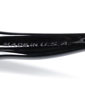 May include: Close-up of a black sunglasses arm. The text "MADE IN U.S.A." is embossed on the arm. The letters "CE" are also visible. The sunglasses appear to be made of a glossy material.