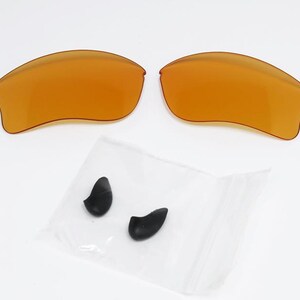 K&ouml;nnte beinhalten: Zwei bernsteinfarbene Ersatz-Sonnenbrillengl&auml;ser und ein kleiner Beutel mit zwei schwarzen Nasenpads. Die Gl&auml;ser sind gew&ouml;lbt und haben eine glatte Oberfl&auml;che. Die Nasenpads sind tropfenf&ouml;rmig.