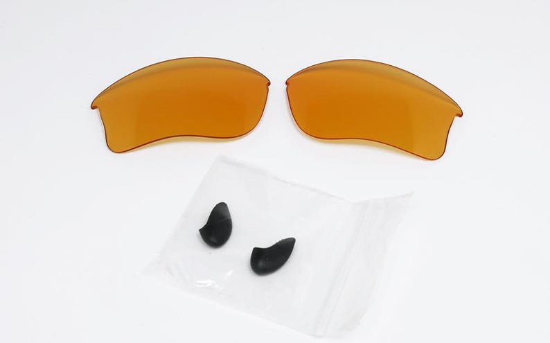 K&ouml;nnte beinhalten: Zwei bernsteinfarbene Ersatz-Sonnenbrillengl&auml;ser und ein kleiner Beutel mit zwei schwarzen Nasenpads. Die Gl&auml;ser sind gew&ouml;lbt und haben eine glatte Oberfl&auml;che. Die Nasenpads sind tropfenf&ouml;rmig.