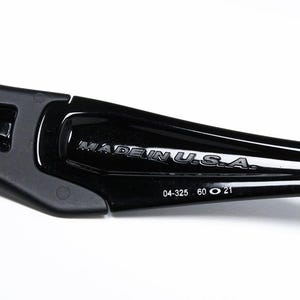 May include: Close-up of a black sunglasses arm with the text "MADE IN U.S.A." in silver lettering. The arm also has the numbers "04-325 60 O 21" printed on it. The sunglasses arm is made of a glossy, durable material.