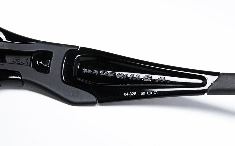 May include: Close-up of a black sunglasses arm with the text "MADE IN U.S.A." in silver lettering. The arm also has the numbers "04-325 60 O 21" printed on it. The sunglasses arm is made of a glossy, durable material.
