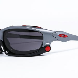 Puede incluir: Gafas de sol grises y negras con detalles rojos. Las gafas tienen un dise&ntilde;o envolvente y una lente grande y oscura.