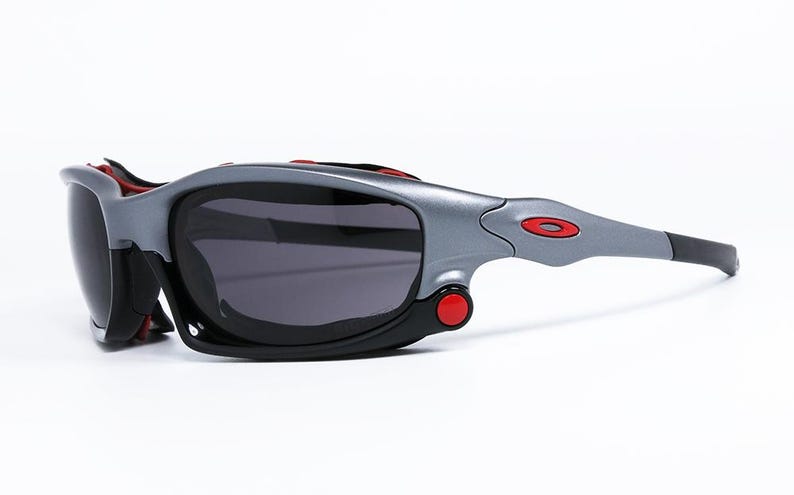 Puede incluir: Gafas de sol grises y negras con detalles rojos. Las gafas tienen un dise&ntilde;o envolvente y una lente grande y oscura.