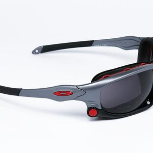 Puede incluir: Gafas de sol grises y negras con detalles rojos. Las gafas tienen un dise&ntilde;o envolvente y lentes grises oscuras.