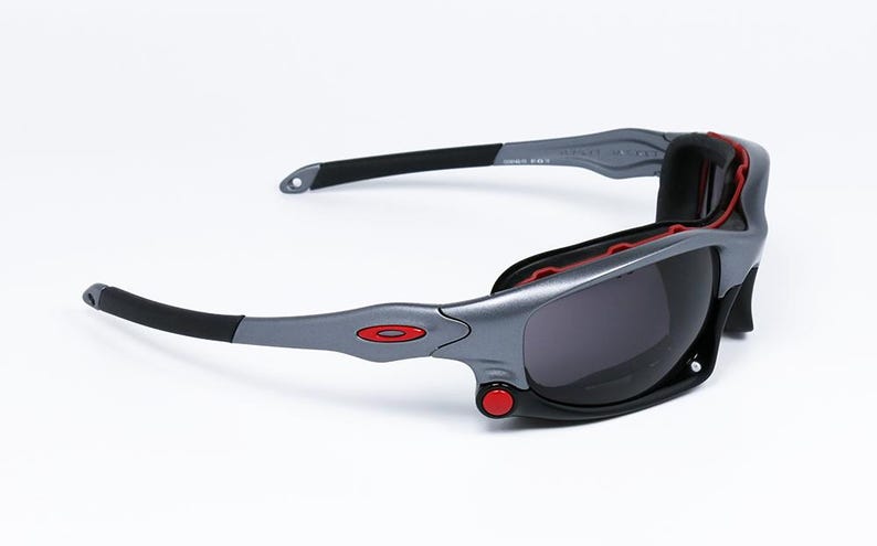 Puede incluir: Gafas de sol grises y negras con detalles rojos. Las gafas tienen un dise&ntilde;o envolvente y lentes grises oscuras.