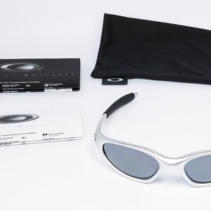 May include: Silver-framed sunglasses with dark lenses, accompanied by a black drawstring pouch, a microfiber cloth, and informational booklets. The sunglasses have a sleek, modern design. The accessories are neatly arranged on a white surface.