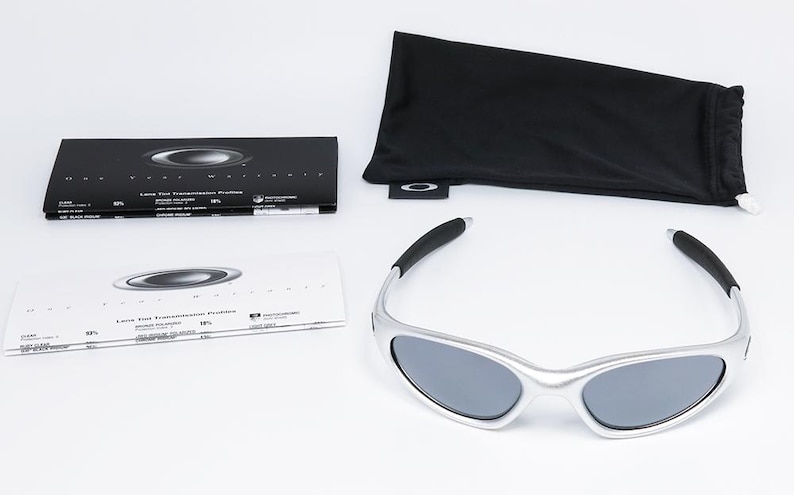 May include: Silver-framed sunglasses with dark lenses, accompanied by a black drawstring pouch, a microfiber cloth, and informational booklets. The sunglasses have a sleek, modern design. The accessories are neatly arranged on a white surface.