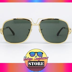 Puede incluir: Gafas de sol doradas con lentes verde oscuro. Las gafas tienen forma cuadrada con doble puente y patillas negras. La imagen incluye un logotipo de Vintage Sunglasses Store.