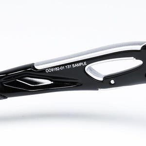 May include: Close-up of the arm of black sunglasses with a silver accent. The text "OO9182-01 131 SAMPLE" is printed on the arm. The sunglasses have a sleek, modern design.