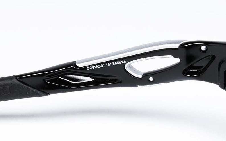 May include: Close-up of the arm of black sunglasses with a silver accent. The text "OO9182-01 131 SAMPLE" is printed on the arm. The sunglasses have a sleek, modern design.