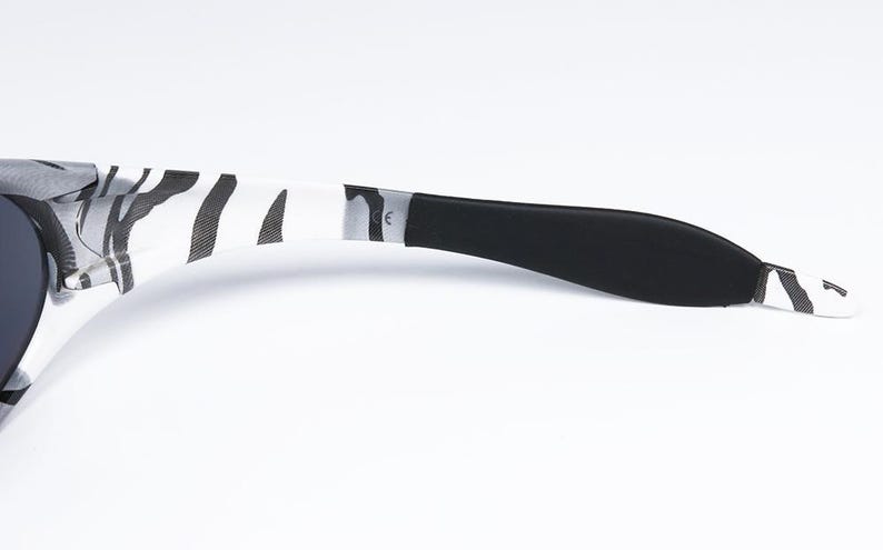 May include: Sunglasses with a black and white zebra print pattern on the arms. The earpiece is black, and the frame is a dark color. The sunglasses are set against a white background.