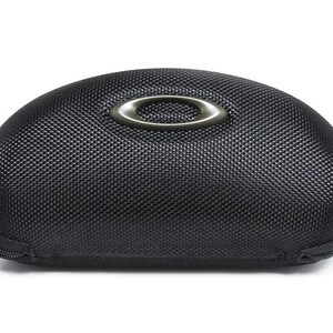 May include: Black, oval-shaped sunglasses case with a textured surface. The case features a silver-colored Oakley logo on the top and a zipper closure. The case is designed to protect eyewear.