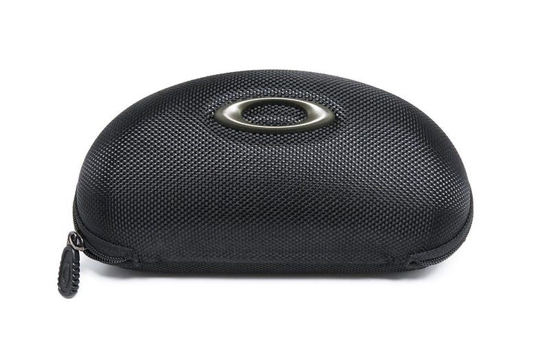 May include: Black, oval-shaped sunglasses case with a textured surface. The case features a silver-colored Oakley logo on the top and a zipper closure. The case is designed to protect eyewear.