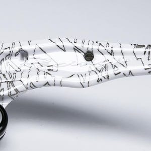 May include: Close-up of a white and black patterned object. The object has a glossy finish and a design of black lines and shapes. A black component with a silver bolt is visible on the left side. The object is on a white background.