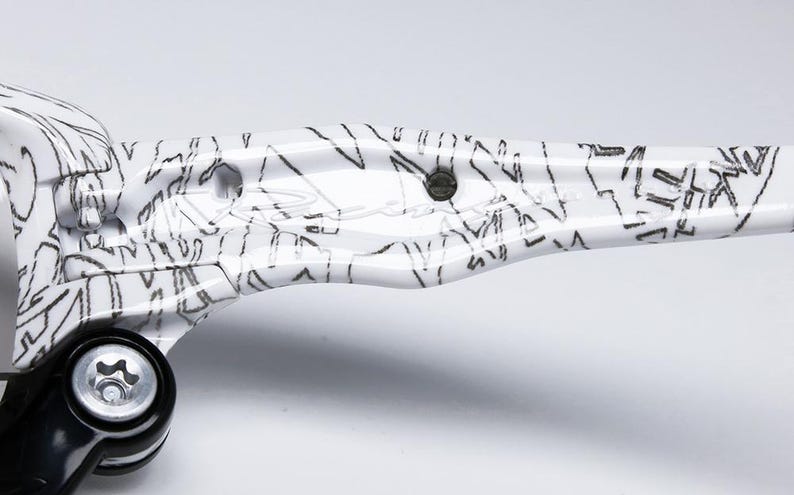 May include: Close-up of a white and black patterned object. The object has a glossy finish and a design of black lines and shapes. A black component with a silver bolt is visible on the left side. The object is on a white background.