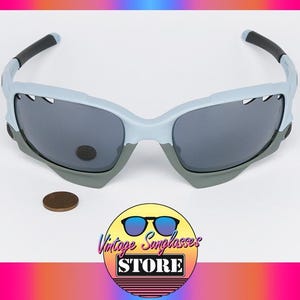 May include: Light blue and gray sunglasses with dark lenses. The sunglasses have a sporty design with black accents on the arms. A penny is next to the sunglasses. The logo for Vintage Sunglasses Store is in the bottom right corner.