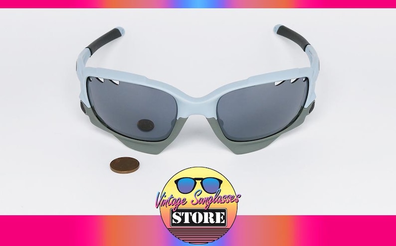 May include: Light blue and gray sunglasses with dark lenses. The sunglasses have a sporty design with black accents on the arms. A penny is next to the sunglasses. The logo for Vintage Sunglasses Store is in the bottom right corner.