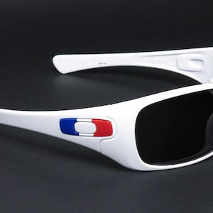 Op de afbeelding: Witte zonnebril met donkere glazen. De monturen hebben een rood, wit en blauw embleem aan de zijkant. De zonnebril heeft een sportief ontwerp en staat tegen een donkere achtergrond.
