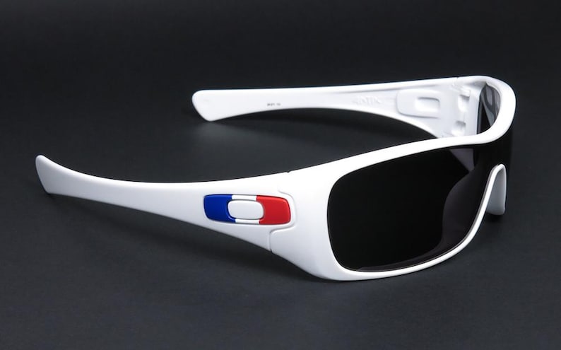 Op de afbeelding: Witte zonnebril met donkere glazen. De monturen hebben een rood, wit en blauw embleem aan de zijkant. De zonnebril heeft een sportief ontwerp en staat tegen een donkere achtergrond.