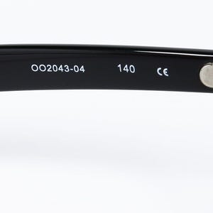 May include: Close-up of the arm of black eyeglasses. The text "OO2043-04" and "140" are printed in white. The hinge is silver. The frame is glossy.