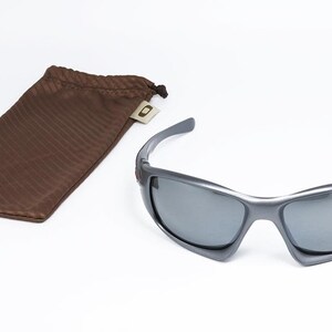 May include: Gray sunglasses with dark lenses and a matching brown drawstring pouch. The sunglasses have a sporty design and are set against a white background. The pouch is made of a textured material.
