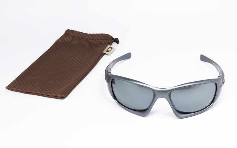 May include: Gray sunglasses with dark lenses and a matching brown drawstring pouch. The sunglasses have a sporty design and are set against a white background. The pouch is made of a textured material.
